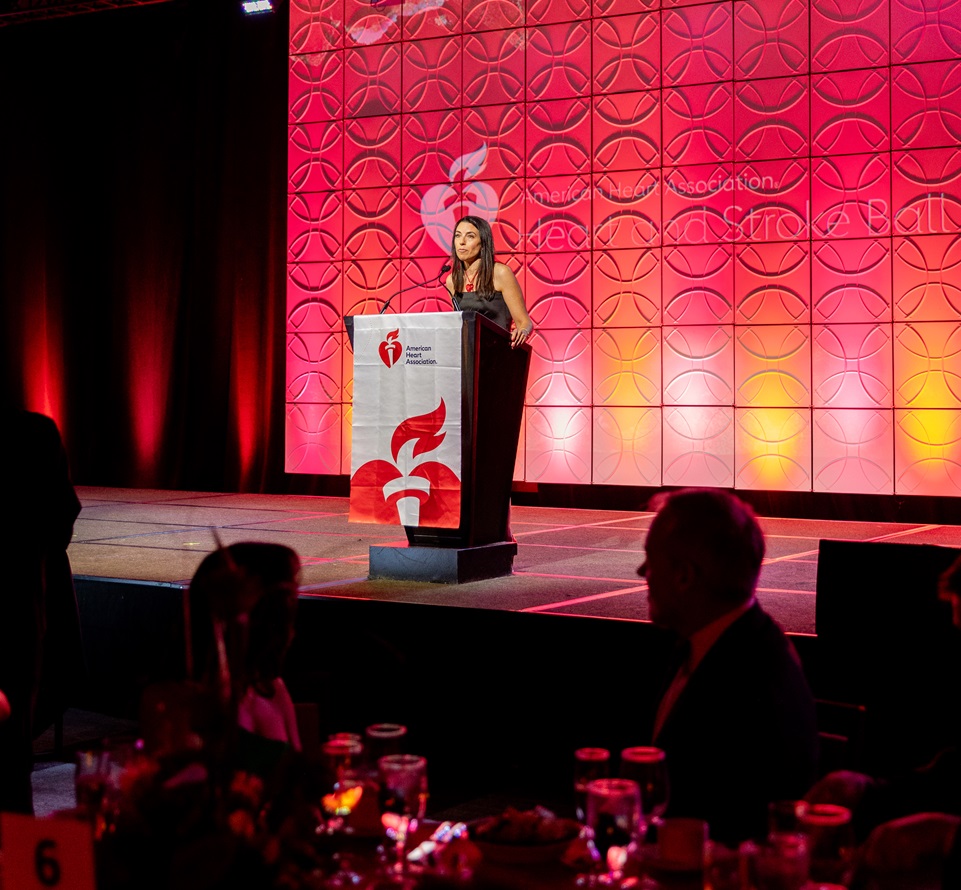 Woman standing at the podium on stage in front of the American Heart Association Heart and Stroke Ball logo projected on the backdrop.